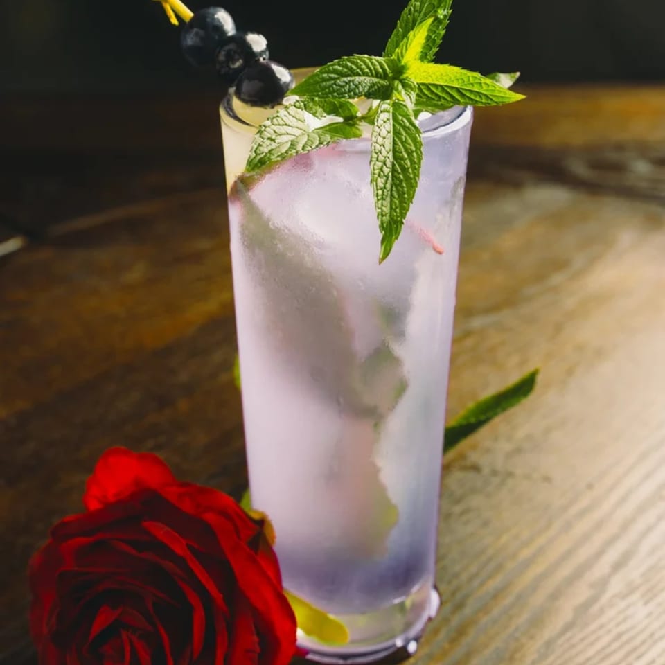 A tall glass of purple cocktail with ice, garnished with blueberries and mint, next to a red rose.