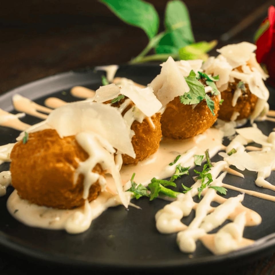 Three golden-brown arancini balls drizzled with white sauce, topped with shaved cheese and cilantro.