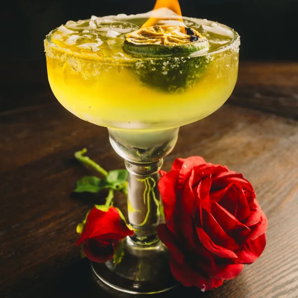 A vibrant yellow margarita with a flaming lime garnish and two red roses on a dark wooden table.