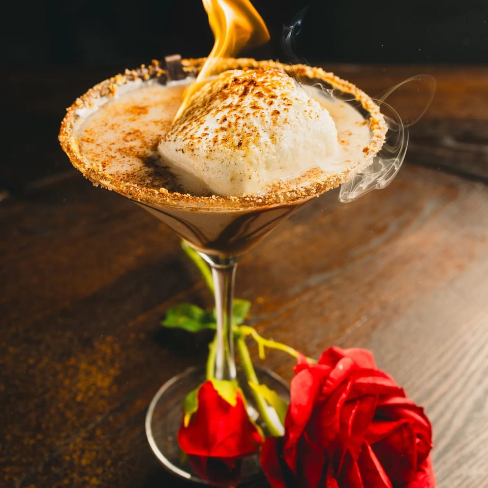 A flaming marshmallow cocktail with a graham cracker rim, garnished with red roses.