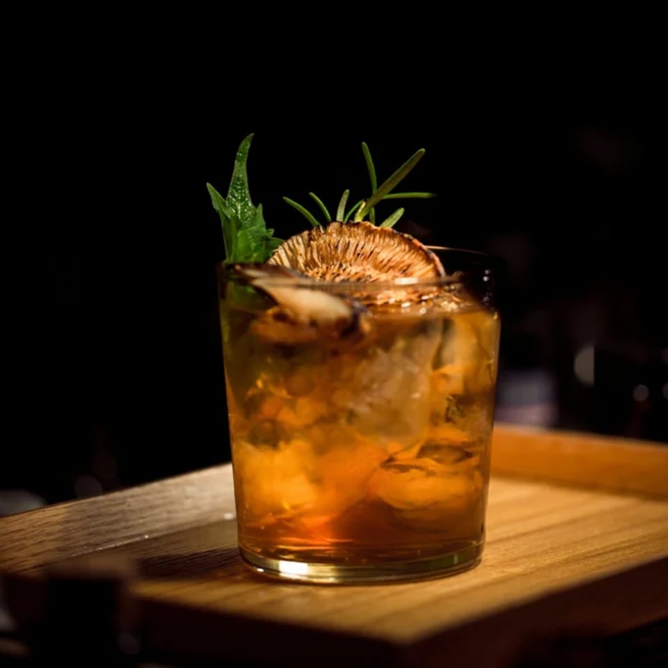 An old fashioned cocktail with ice, garnished with dried citrus and herbs on a wooden tray.
