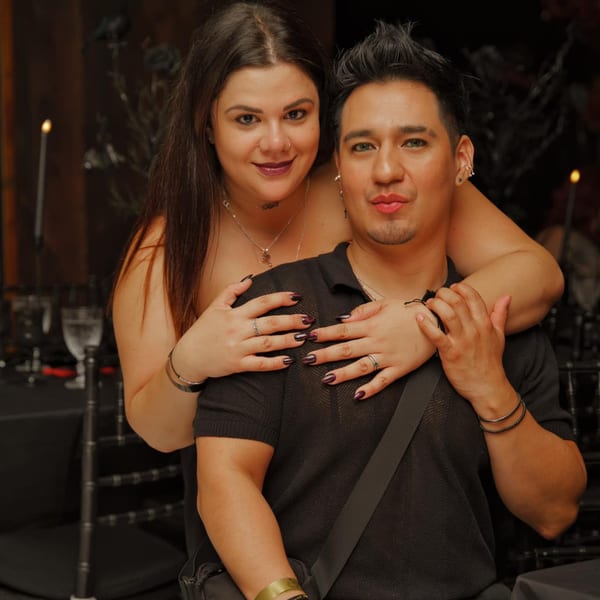A woman with dark hair embraces a man from behind, both smiling at the camera.
