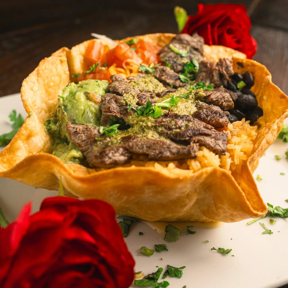 A taco salad in a crispy shell with steak, guacamole, rice, beans, and tomatoes, garnished with red roses.