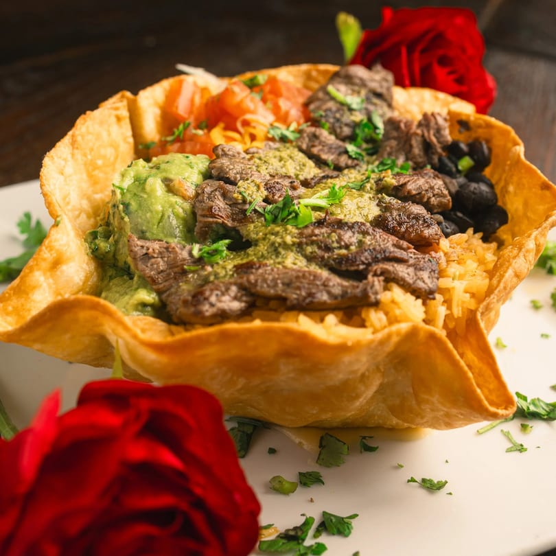 A taco salad in a crispy shell with steak, guacamole, rice, beans, and tomatoes, garnished with red roses.