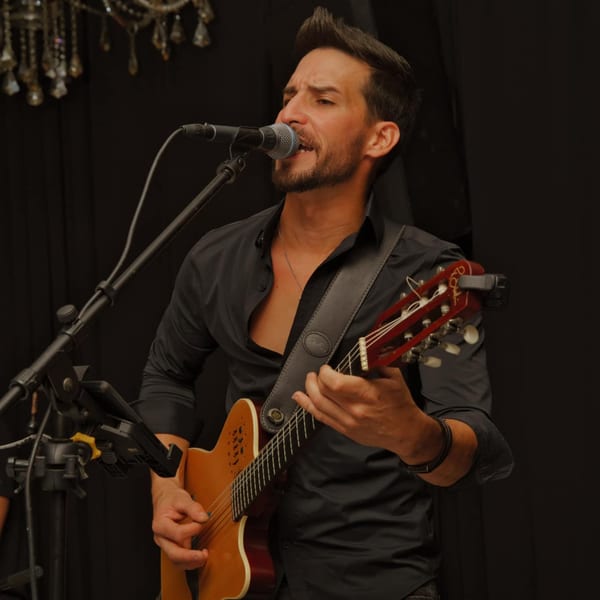 A male musician sings into a microphone while playing an acoustic guitar on a dark stage.