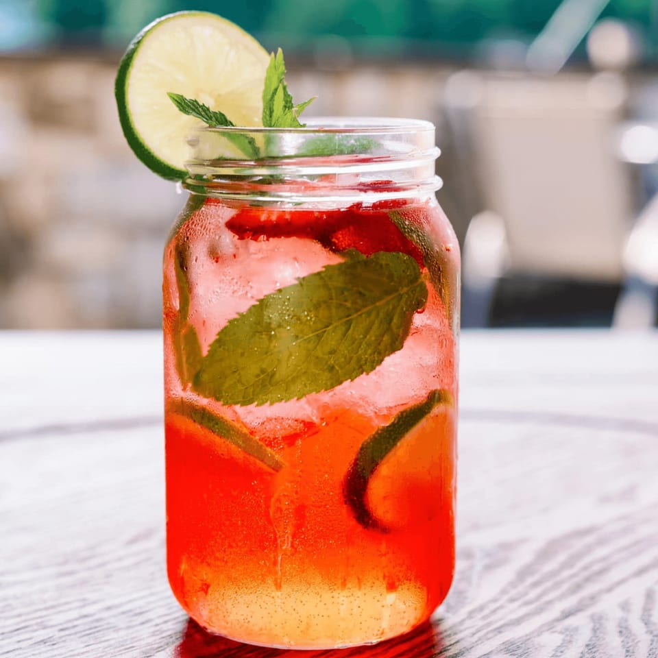 Refreshing red drink in a mason jar with lime, mint, and strawberries on a wooden table.